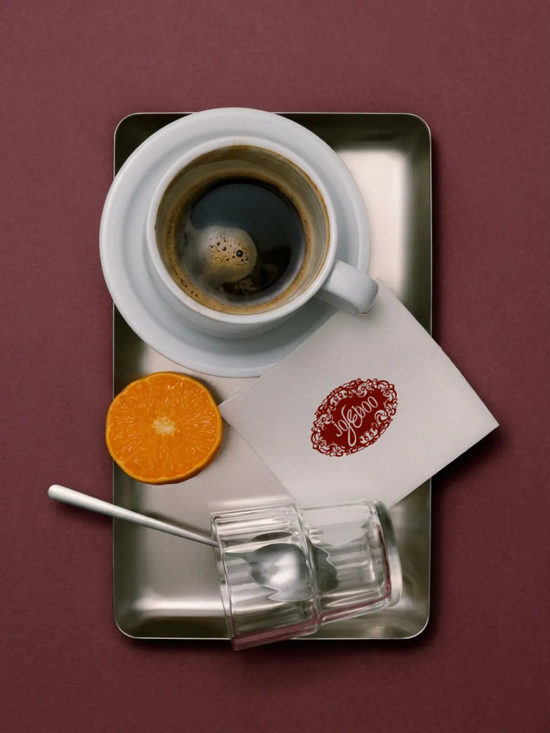 Social post: a steel tray with an espresso, half an orange, a tipped glass and a napkin printed with the lace seal, on burgundy