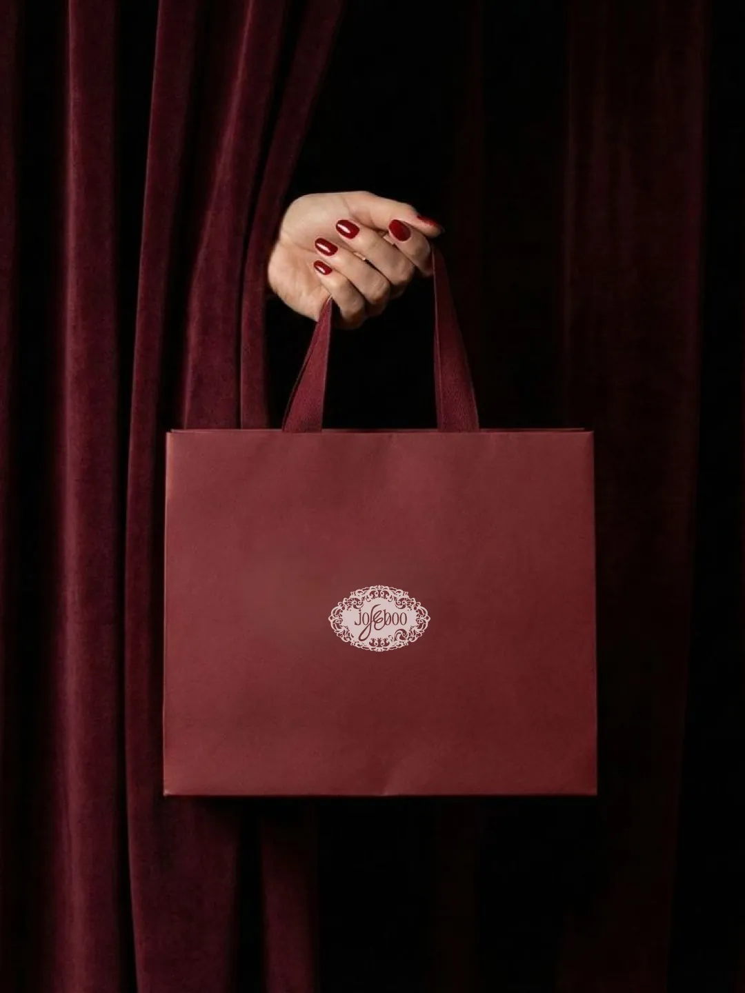 Social post: a hand with red nails reaching through a burgundy velvet curtain, holding a burgundy shopping bag with the lace seal