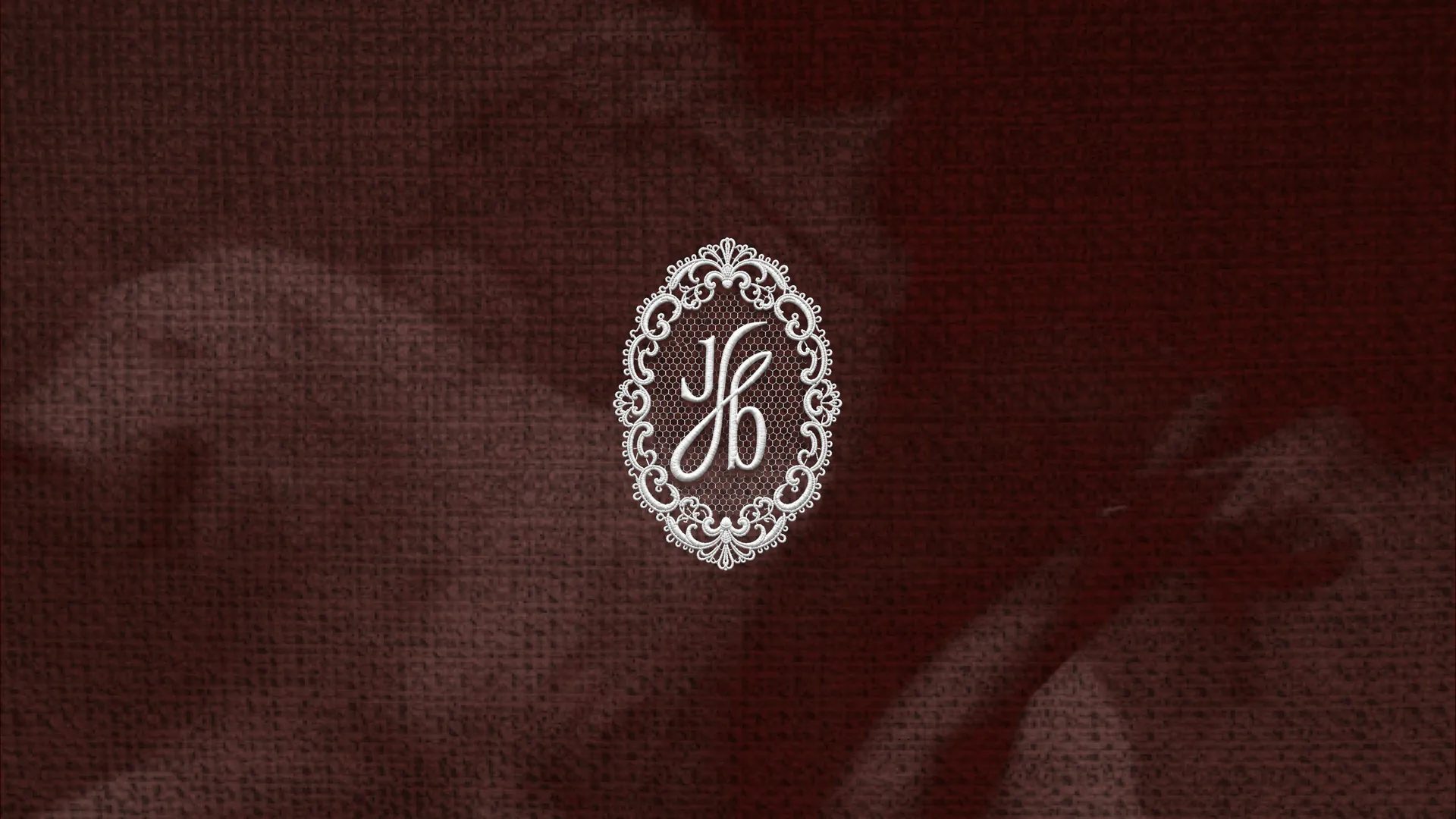 The Jb monogram inside an oval lace ring, rendered as white embroidery on dark red textured cloth