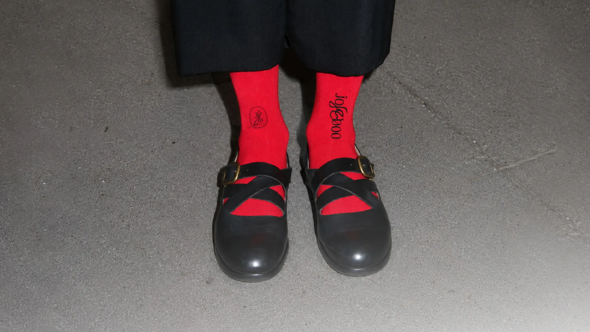 Red ribbed socks and black Mary Jane shoes under a direct flash on grey concrete, the jojeeboo wordmark running down one leg and the Jb in a thin oval on the other